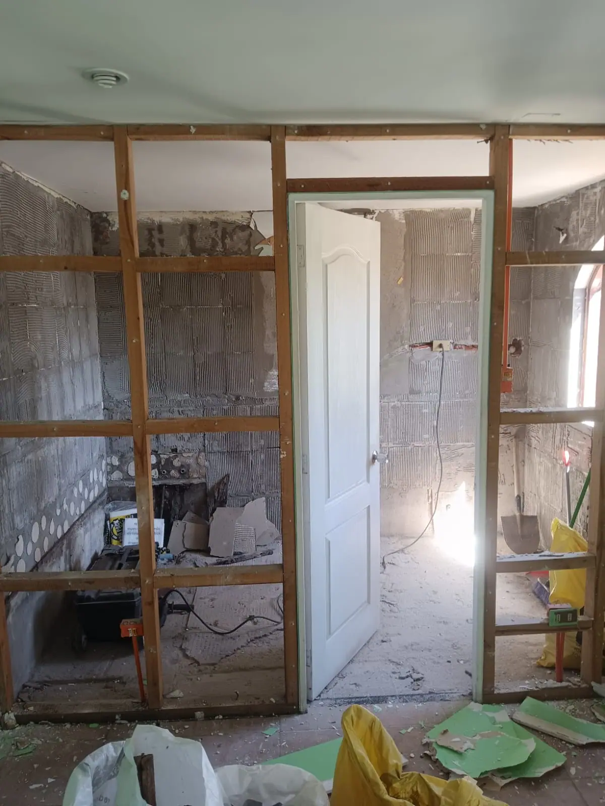 Baño desarmado hasta la estructura de madera, con la puerta antigua todavía montada y escombro en el suelo.