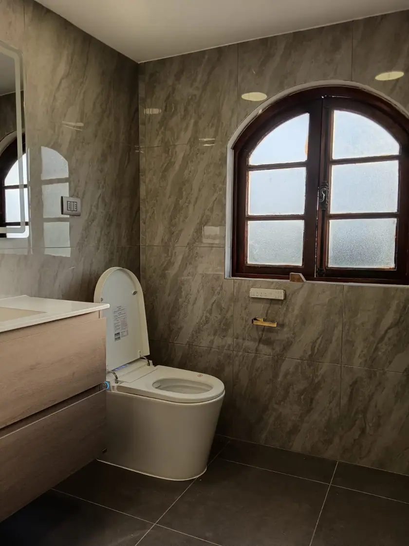 Baño terminado con revestimiento porcelánico de gran formato, mueble de madera bajo el lavamanos y la ventana de arco conservada.