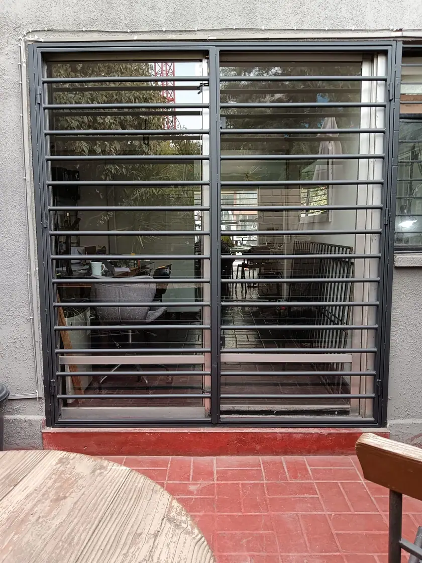 Reja instalada y pintada en gris oscuro sobre una puerta corredera de vidrio, vista desde el patio.