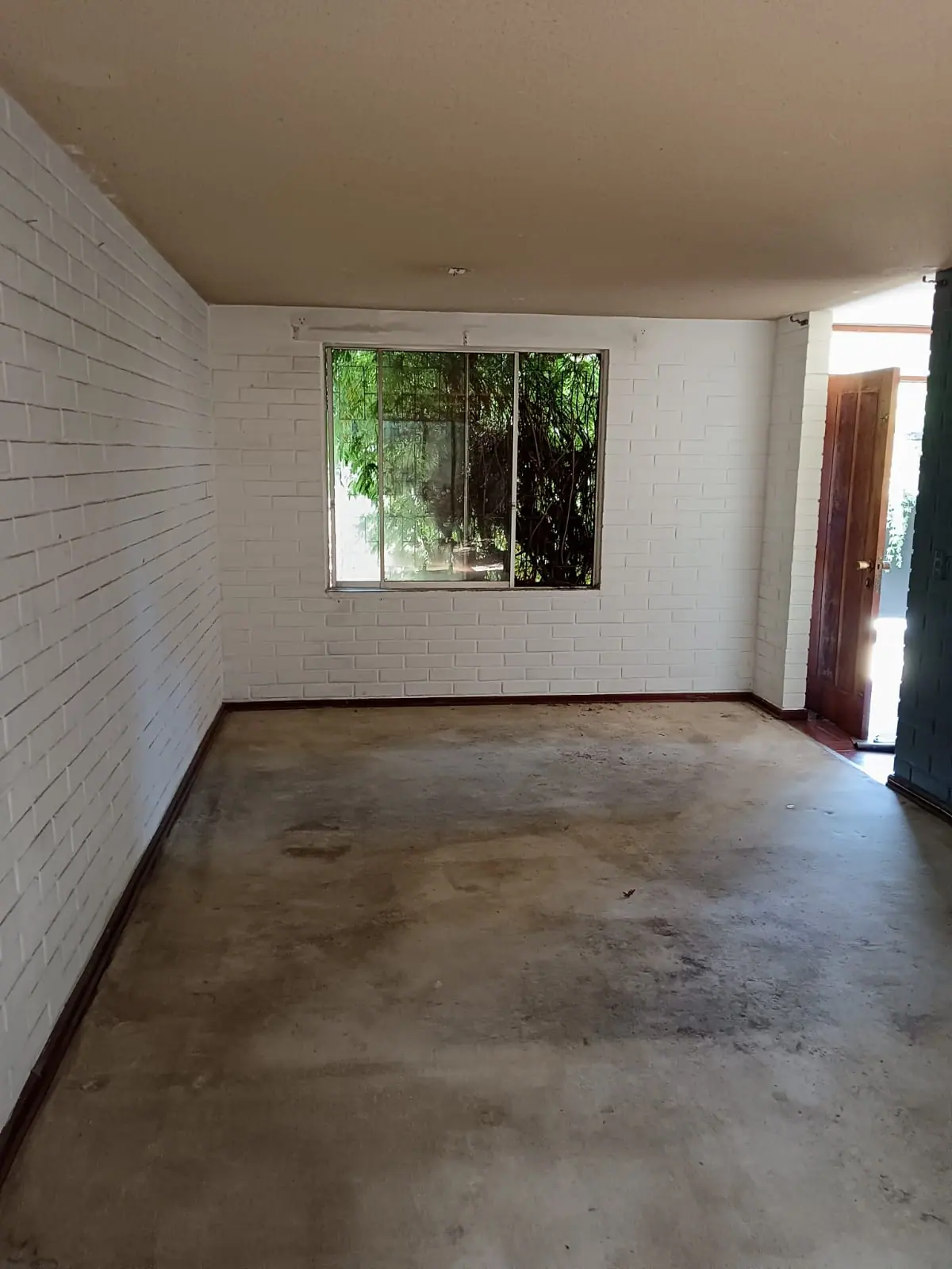 Recinto vacío con muros de albañilería pintados de blanco, piso de hormigón sin terminación y una ventana con reja.