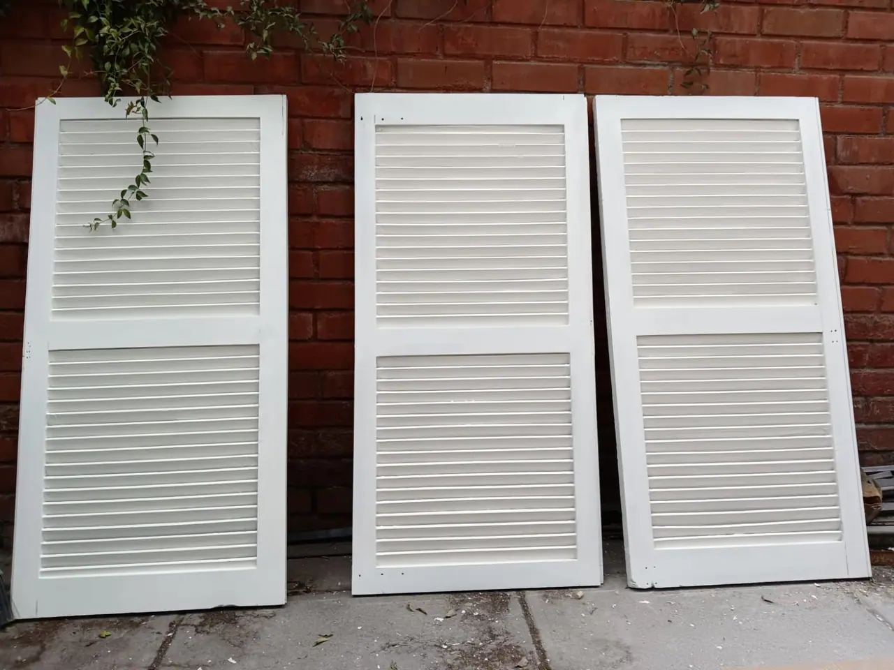 Tres hojas de persiana terminadas y pintadas de blanco, apoyadas contra un muro de ladrillo.