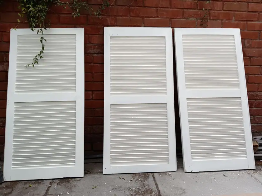 Tres hojas de persiana terminadas y pintadas de blanco, apoyadas contra un muro de ladrillo.